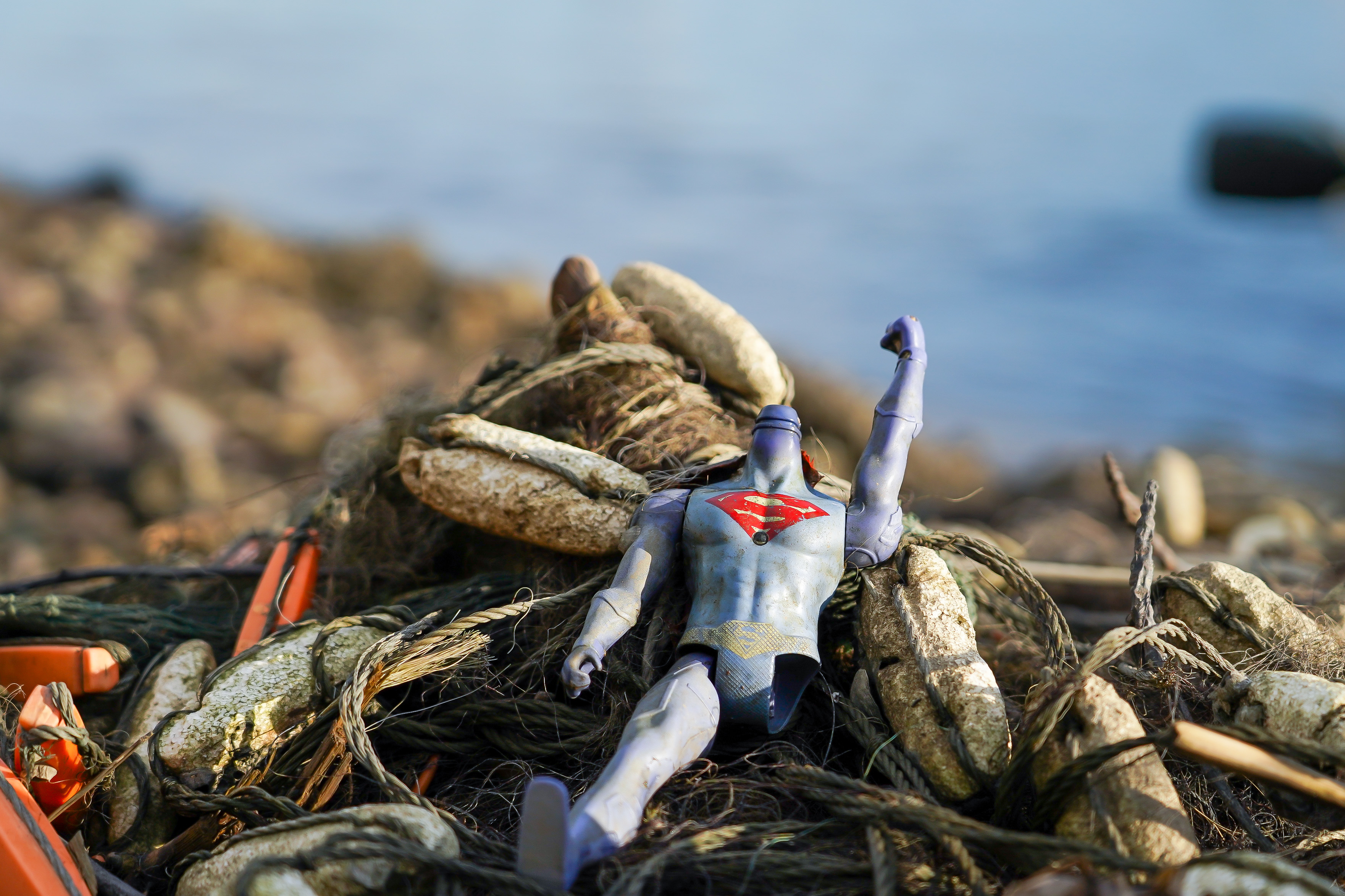 En ødelagt superhelt-leke ligger blant tau og søppel ved vannkanten.