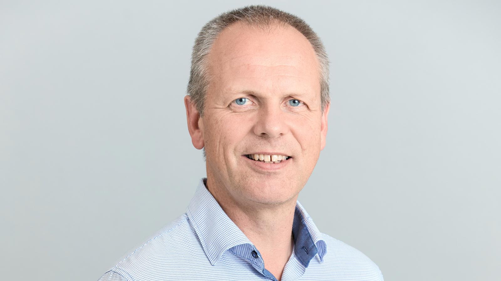 En mann med kort, lyst hår står foran en ensfarget, lys bakgrunn. Han har på seg en lyseblå skjorte med små ruter.