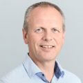 En mann med kort, lyst hår står foran en ensfarget, lys bakgrunn. Han har på seg en lyseblå skjorte med små ruter.