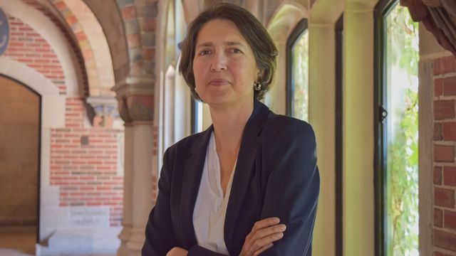 A woman with medium-length hair stands with her arms crossed, wearing a dark blazer over a light shirt, in a sunlit hallway with brick arches and tall windows.