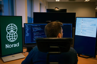 A man is sitting at a desk in an office, seen from behind, working on three computer monitors; one displays the Norad logo, another shows lines of programming code, and the third displays a document or email interface.