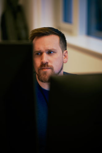 A young man sits at a desk, partially obscured by computer monitors, with windows in the background.