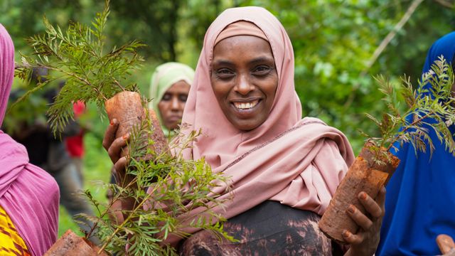 Kvinne med rosa sjal smiler til kameraet. Hun holder planter i begge hendene.