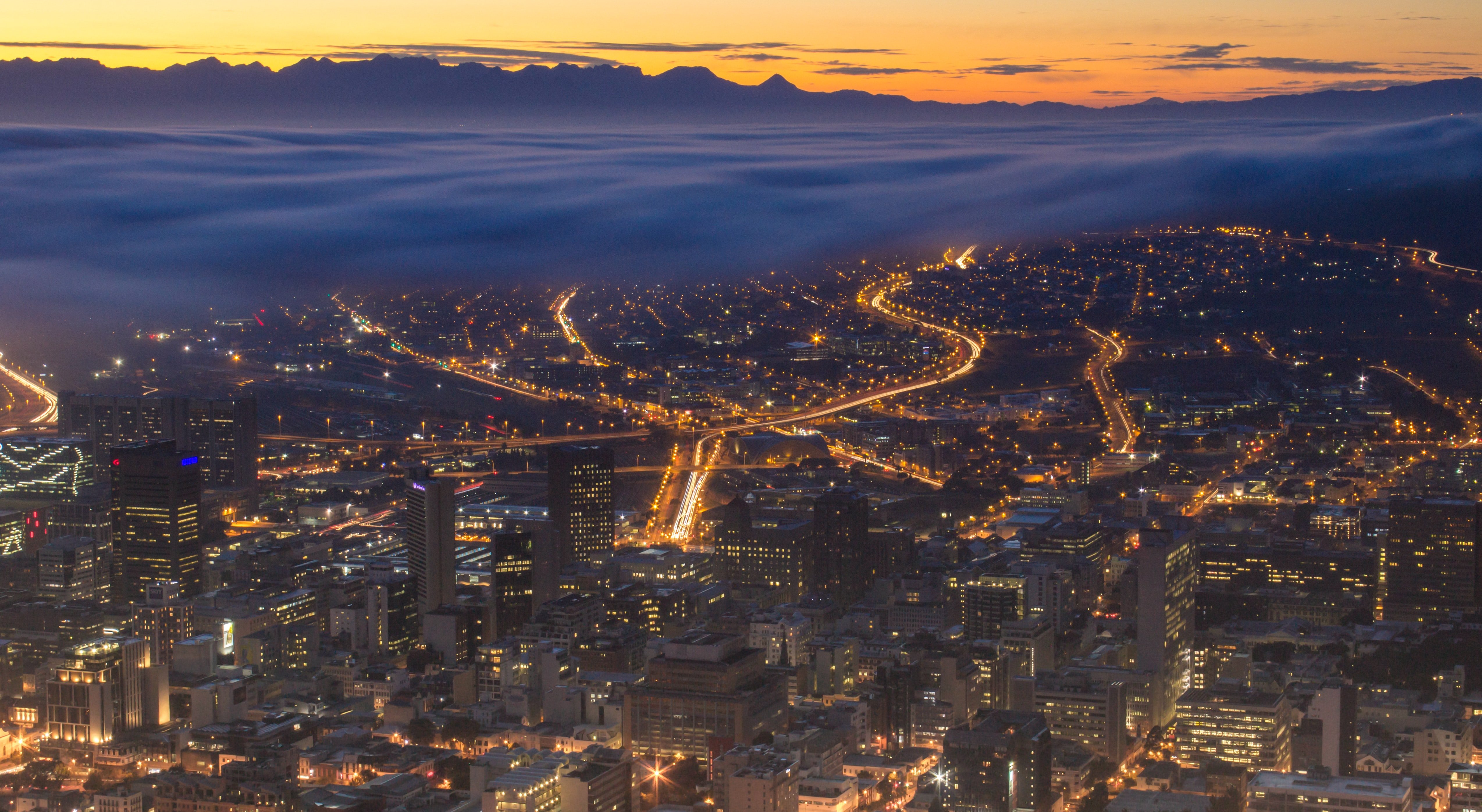 A cityscape is captured at dusk, with city lights illuminating the buildings and winding roads below. Layers of low-lying clouds or mist stretch across the middle of the image, partially covering the outskirts of the city. In the background, mountain silhouettes are visible against a dramatic orange and yellow sunset sky. The overall scene creates a striking contrast between the glowing city and the natural landscape.