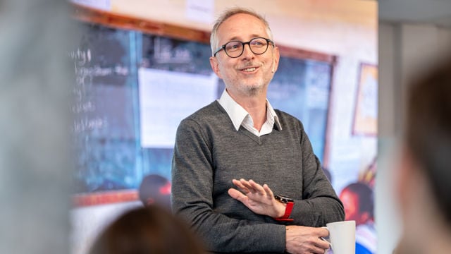 Bildet viser en smilende Bård Vegar SolhjellHan har på seg en grå genser over en hvit skjorte og holder en kaffekopp i den ene hånden.