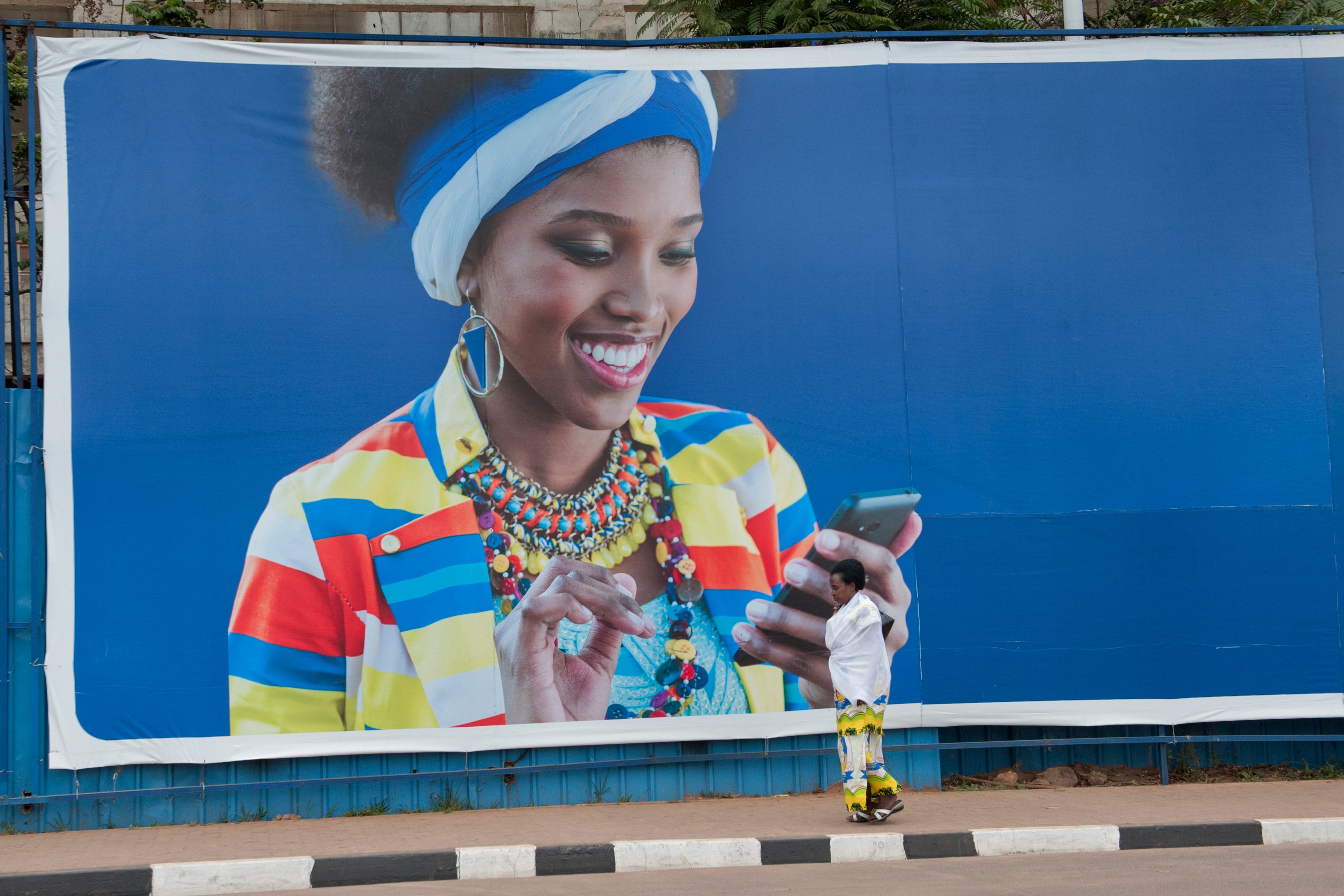 A group of people are working together outdoors on a construction project, possibly building a foundation with stones and earth. In the foreground, two women stand observing the work; one is dressed in a pink headscarf and patterned skirt, while the other carries a child on her back secured with a colorful cloth. Several men are actively involved in the construction, using tools to move and position rocks. The setting is rural, with green grass, scattered stones, and hills visible in the background.