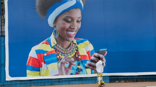 A group of people are working together outdoors on a construction project, possibly building a foundation with stones and earth. In the foreground, two women stand observing the work; one is dressed in a pink headscarf and patterned skirt, while the other carries a child on her back secured with a colorful cloth. Several men are actively involved in the construction, using tools to move and position rocks. The setting is rural, with green grass, scattered stones, and hills visible in the background.