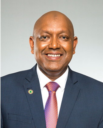 Picture of Dr. Yero Baldeh wearing a dark blue suit, a white shirt, and a colorful red and blue patterned tie. He is standing against a neutral, light background. On his lapel, he has a round pin with a green and yellow design.