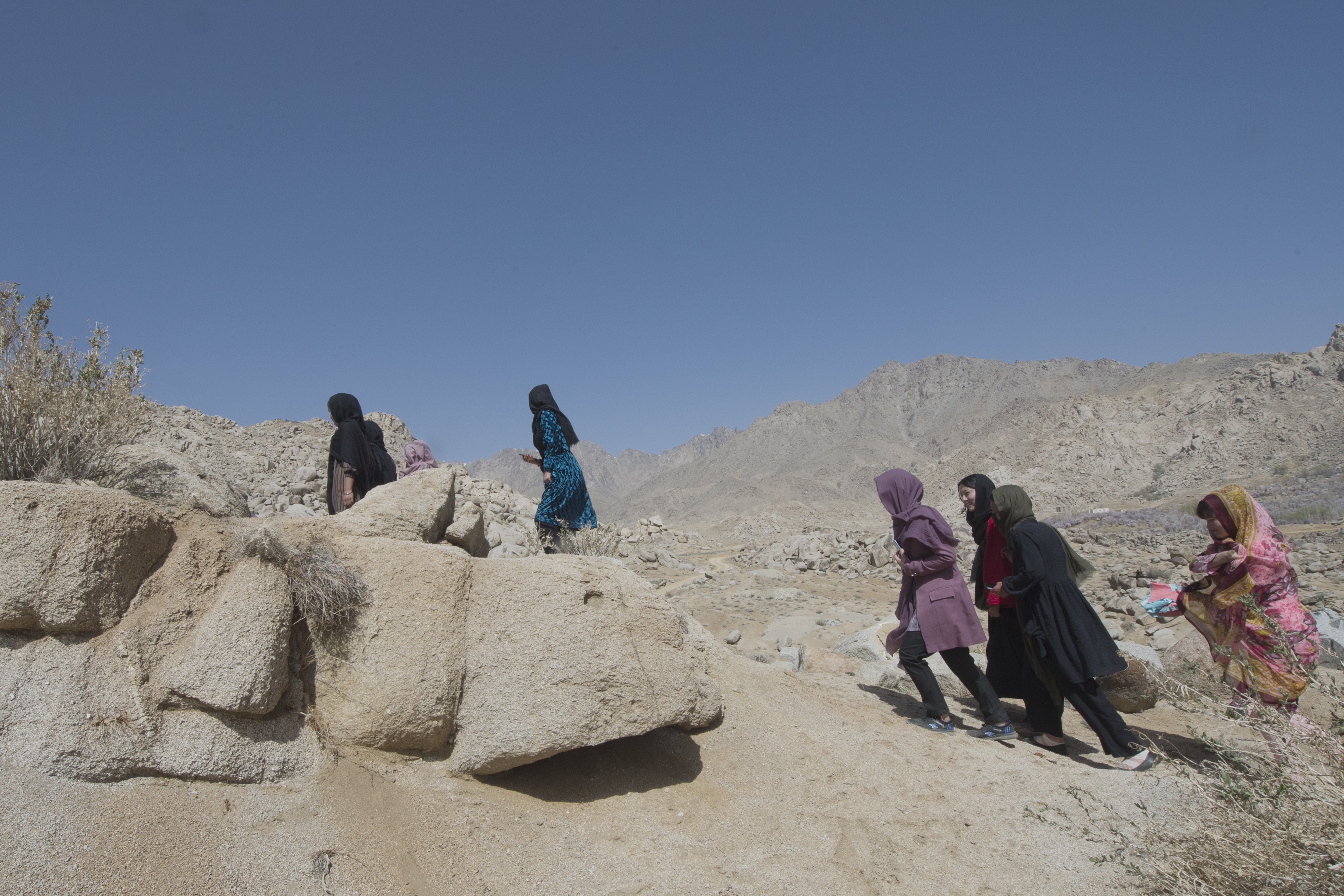Young people walk up a rocky slope in a dry landscape under a clear blue sky. Some are close to large rocks in the foreground, while others continue along the path uphill.