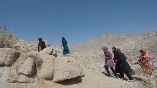 Young people walk up a rocky slope in a dry landscape under a clear blue sky. Some are close to large rocks in the foreground, while others continue along the path uphill.