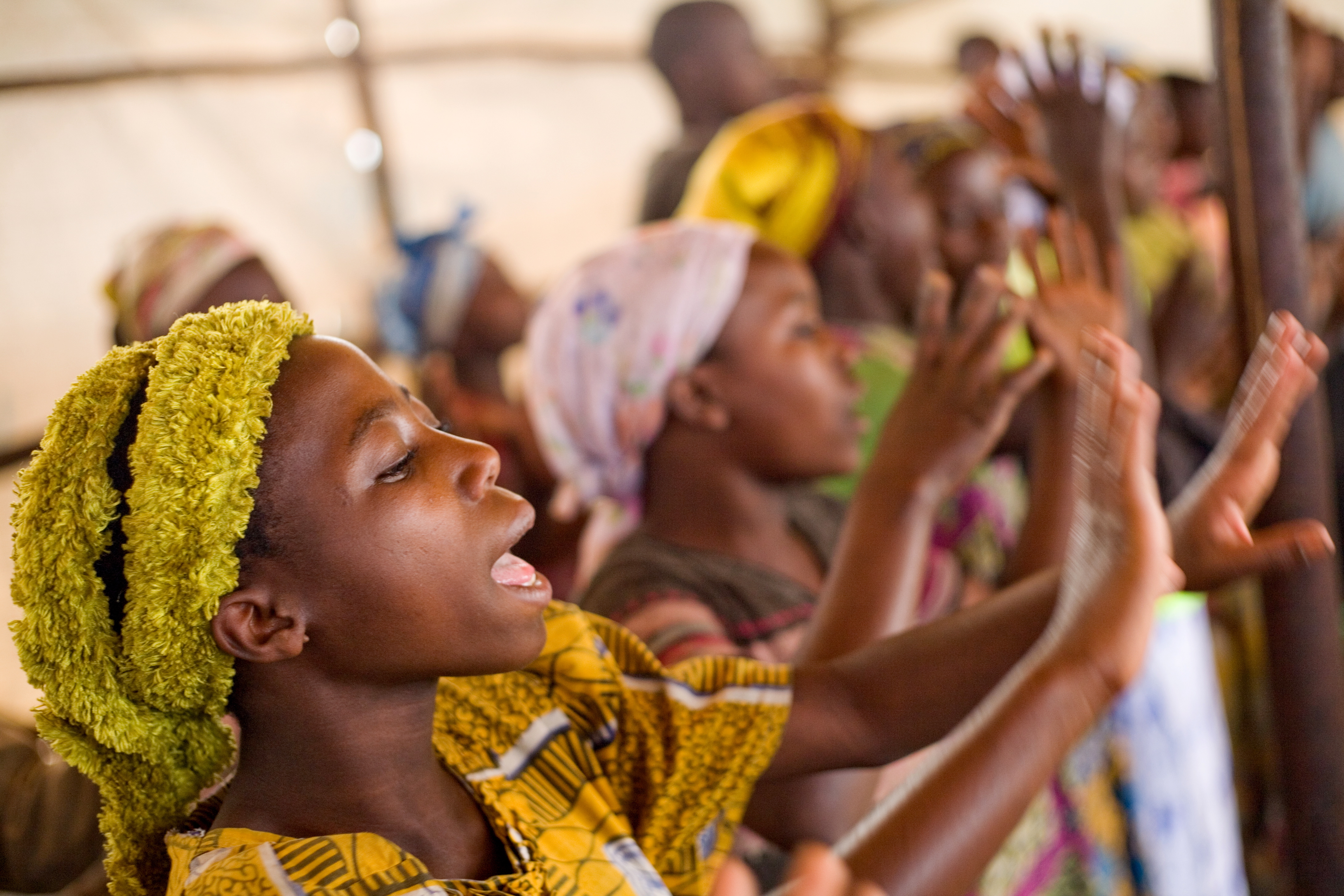 En gruppe unge jenter kledd i fargerike klær står i en rekke og klapper hendene, innendørs.