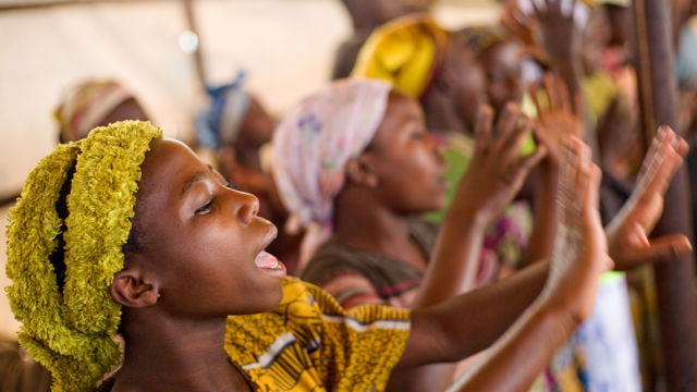 En gruppe unge jenter kledd i fargerike klær står i en rekke og klapper hendene, innendørs.
