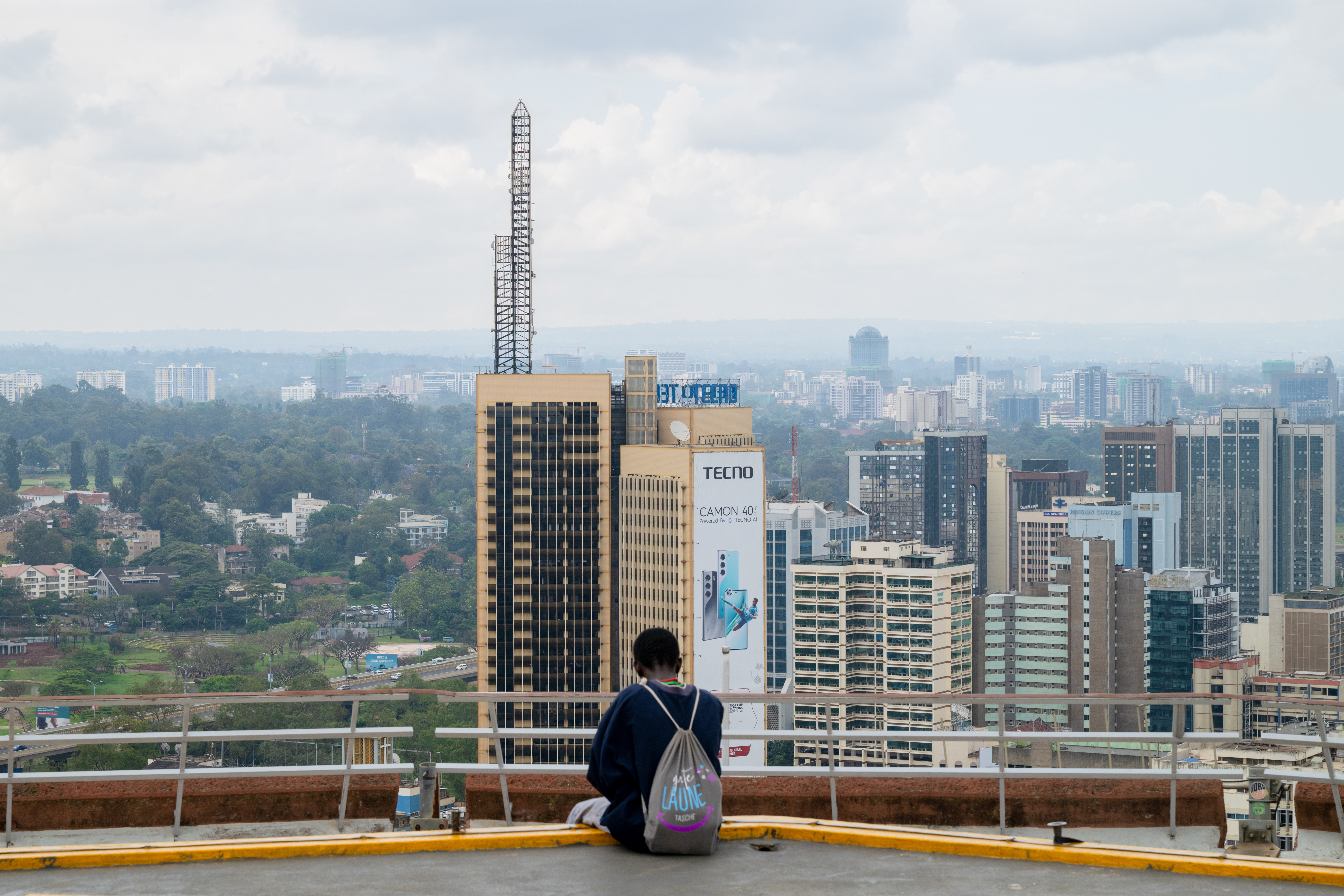 Man with backpack sitting on a rooftop overlooking skyscrapers in a huge city. The sky is cloudy and there are green trees in the background.