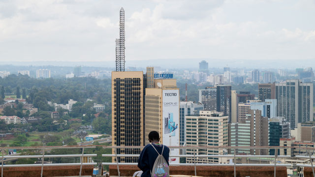 Man with backpack sitting on a rooftop overlooking skyscrapers in a huge city. The sky is cloudy and there are green trees in the background.