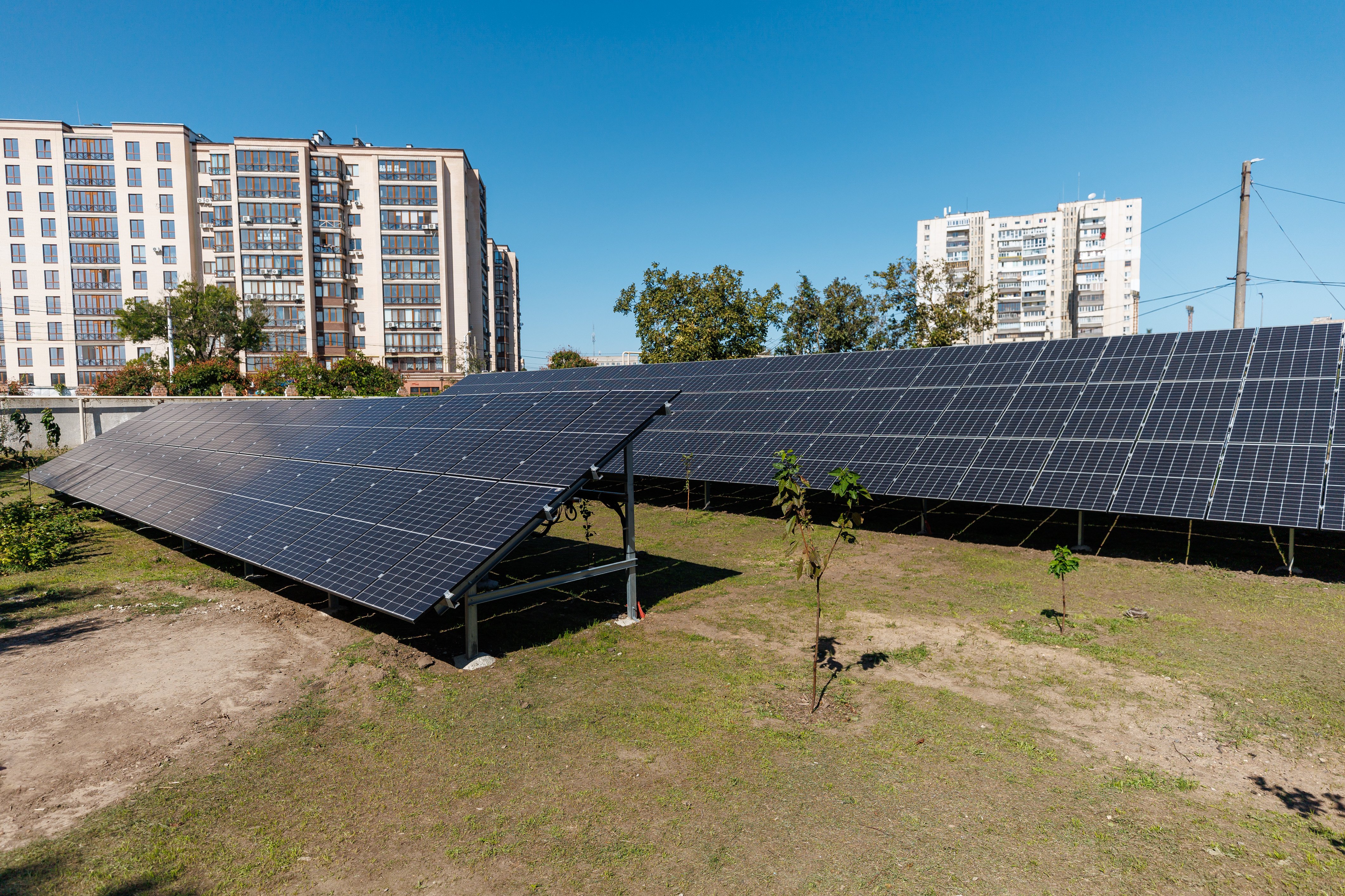 To rader med solcellepaneler står plassert på en gressplen foran flere høyblokker i et byområde.