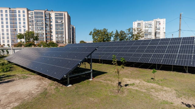 Two rows of solar panels are installed on a grassy area in front of several apartment buildings in an urban setting.
