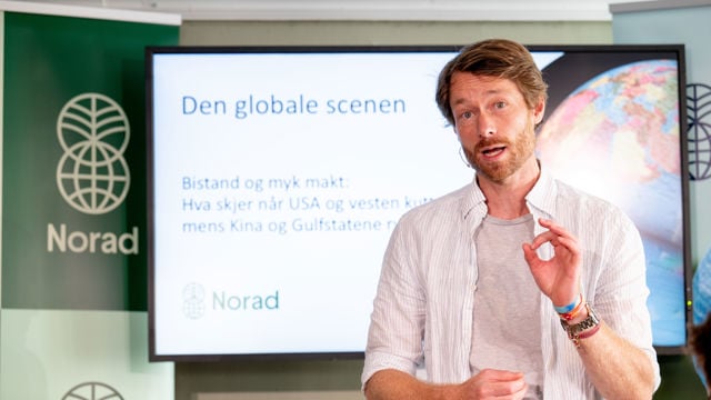 En ung mann står foran en skjerm med Norad-logo og presentasjonstekst om bistand og internasjonale maktforhold.