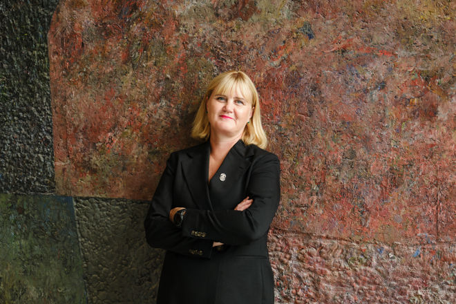 En voksen kvinne med lyst hår står med armene i kors foran en fargerik vegg med grov tekstur.