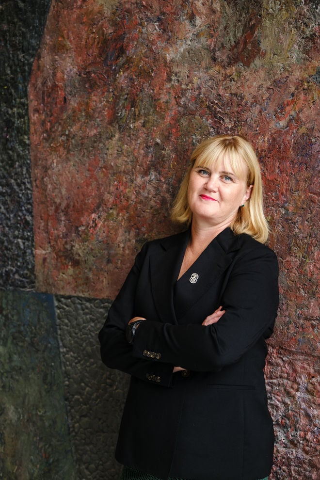 Bildet viser en kvinne med skulderlangt blondt hår som står med armene i kors foran en vegg med et kunstnerisk, teksturert og fargerikt mønster. Hun har på seg en svart blazer med en brosje på jakkeslaget og et mørkt armbåndsur. Stemningen er formell og profesjonell.