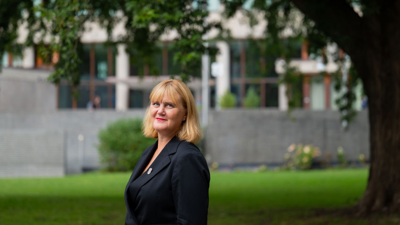 A woman with blonde hair and a dark blazer is standing on a lawn in front of a building and a large tree.