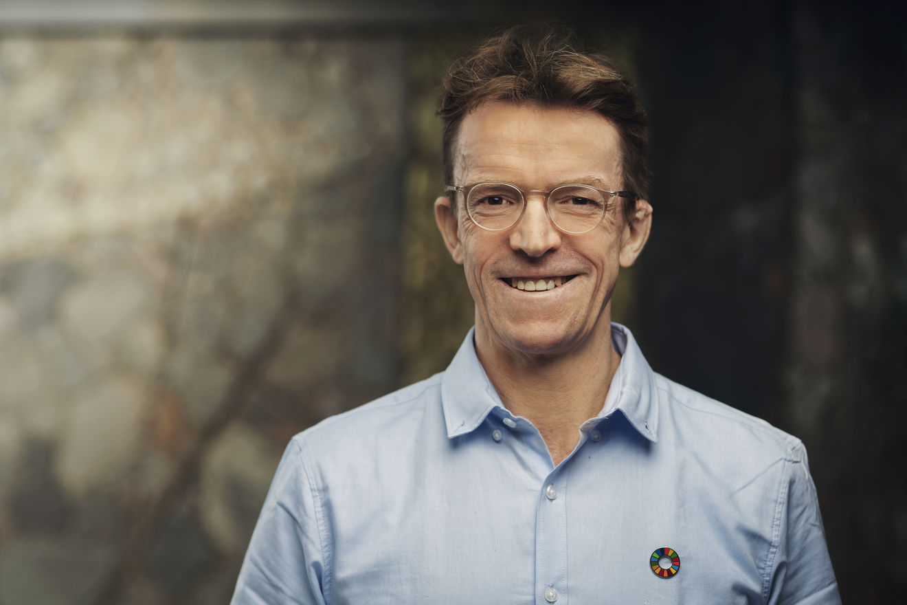 A man wearing a light blue shirt with a pin featuring the Sustainable Development Goals symbol stands indoors in front of a blurred background.