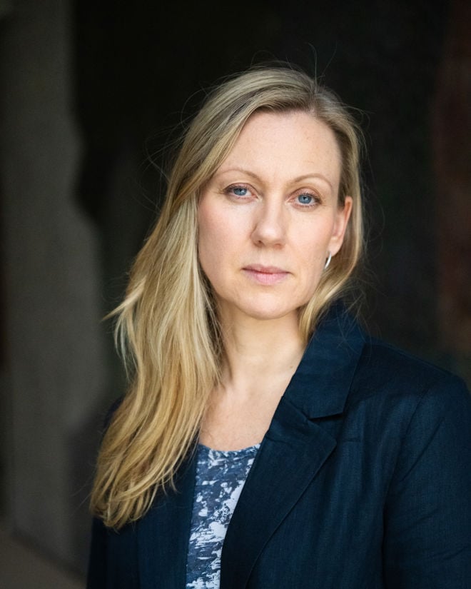 En blond kvinne i mørk blazer og blå topp står foran en mørk bakgrunn og ser mot kameraet.