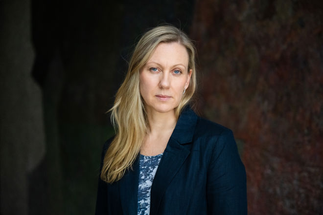 A young woman with long blonde hair is standing indoors, wearing a dark jacket.