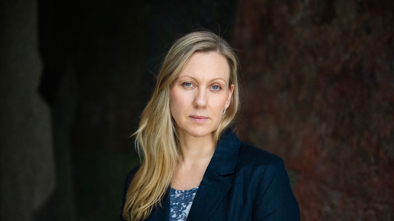 En voksen kvinne med langt lyst hår står foran en mørk bakgrunn, iført blå blazer og mønstret topp.
