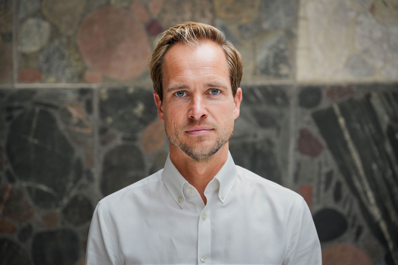 En ung mann med lyst hår og hvit skjorte står foran en vegg med ulike steinmønstre.
