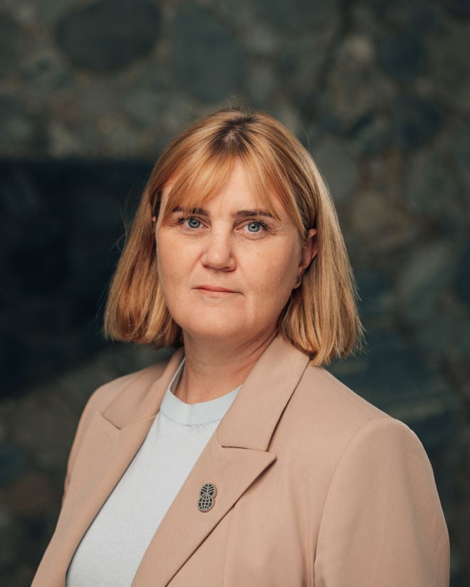 Bildet viser en voksen kvinne med lyst, skulderlangt hår, iført lys blazer og hvit topp, fotografert foran en mørk bakgrunn.
