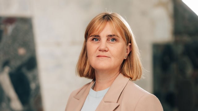 Bildet viser en voksen kvinne med lyst, skulderlangt hår. Hun har på seg en lys blazer og en lyseblå topp, og står foran en uklar bakgrunn innendørs.