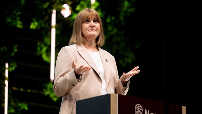 En kvinne med lyst hår står bak en talerstol og holder en presentasjon innendørs.