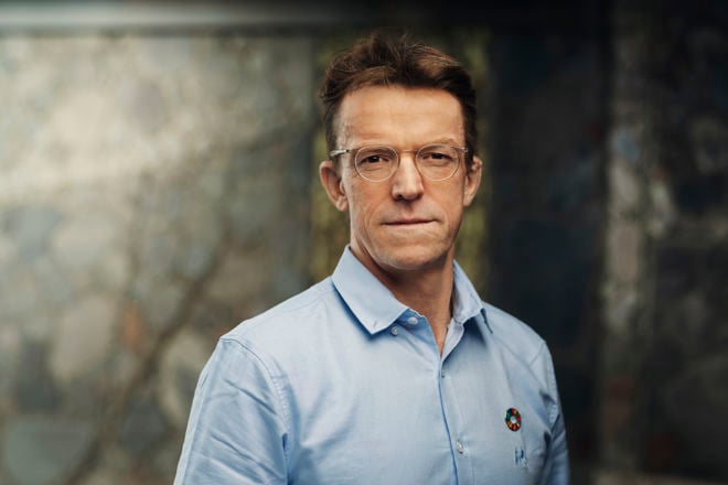 En mann med brunt hår iført lyseblå skjorte med bærekraftsmål-pin, står foran en steinvegg.