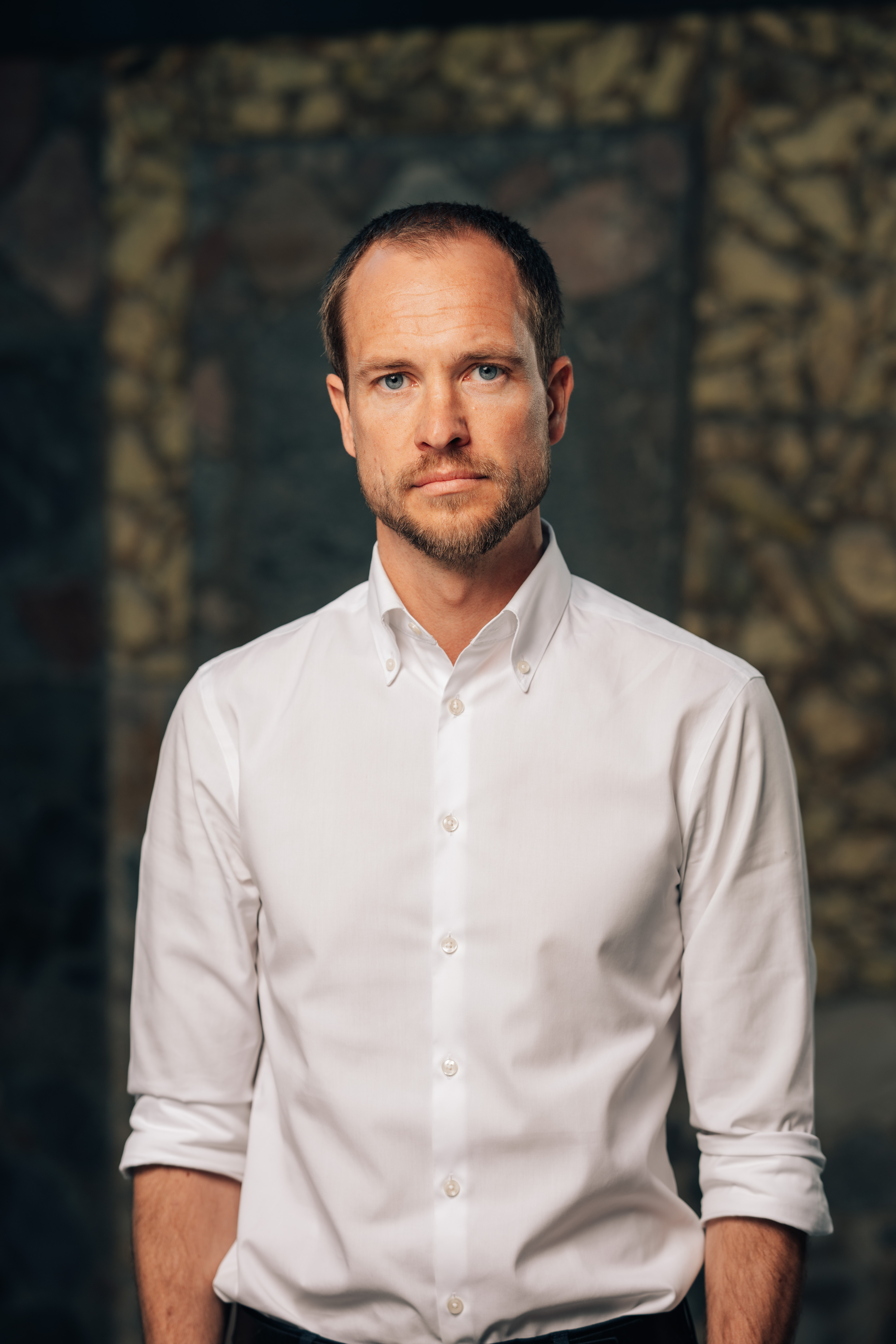 En voksen mann står foran en steinmur, iført hvit skjorte med oppbrettede ermer.