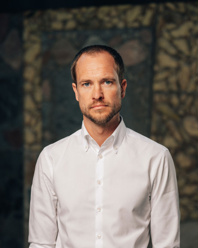 En voksen mann står foran en steinmur, iført hvit skjorte med oppbrettede ermer.