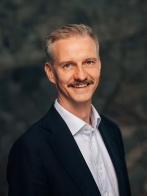 En voksen mann med lyst, kort hår iført hvit skjorte og mørk dressjakke, står foran en mørk steinbakgrunn.