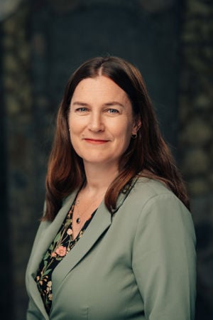 En voksen kvinne med langt brunt hår står foran en mørk bakgrunn, iført en lysegrønn blazer og blomstret topp.