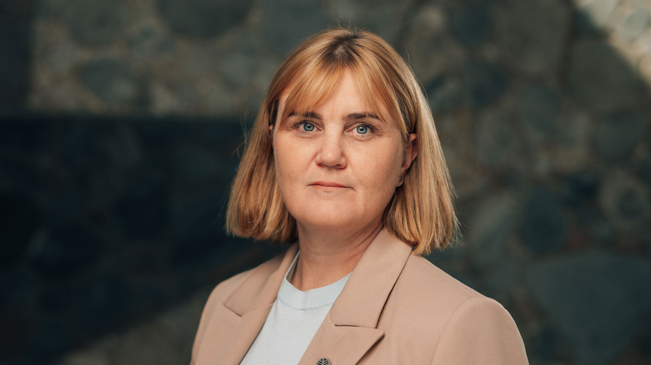 Portrett av en voksen kvinne i lys blazer og lys topp foran en mørk steinvegg.