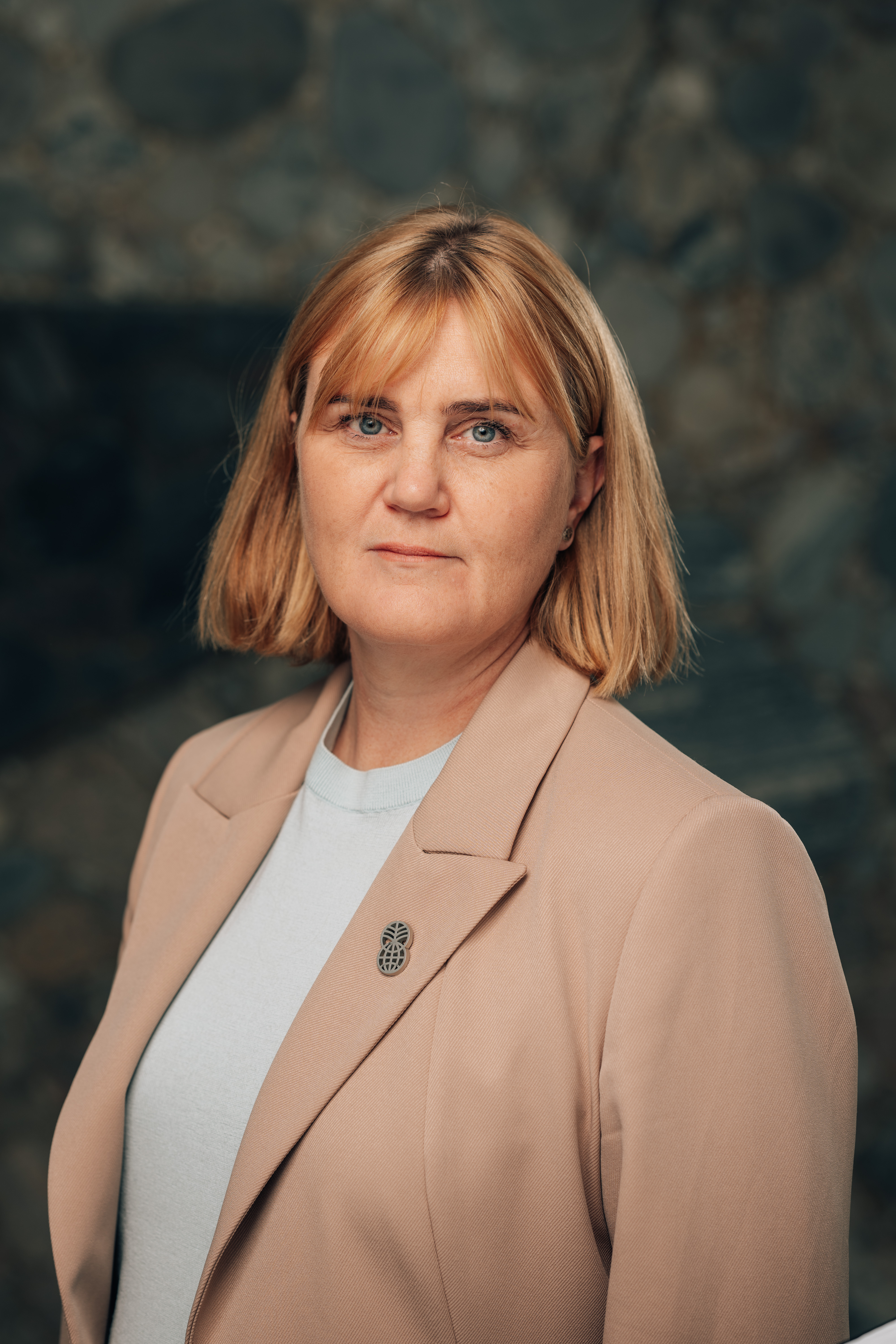 Portrett av en voksen kvinne med lyst, kort hår, iført lys topp og beige blazer, foran en mørk bakgrunn.