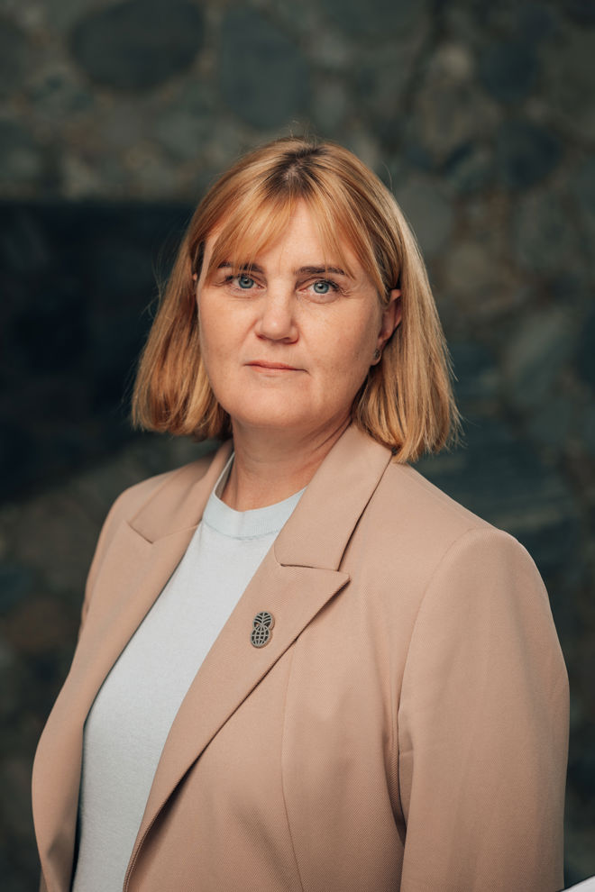 Portrett av en voksen kvinne med lyst, kort hår, iført lys topp og beige blazer, foran en mørk bakgrunn.