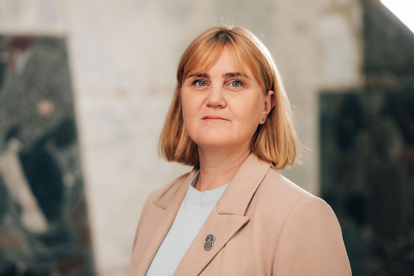 Portrett av en voksen kvinne med kort lyst hår, iført lys overdel og beige blazer, stående innendørs foran en uskarp bakgrunn.