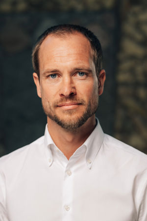En voksen mann med kort mørkt hår står foran en mørk bakgrunn, iført en hvit skjorte.