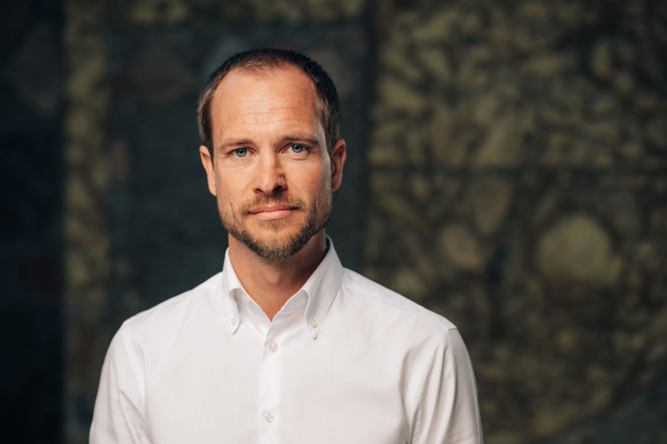 Portrett av en voksen mann i hvit skjorte, fotografert forfra mot en mørk, mønstret bakgrunn.