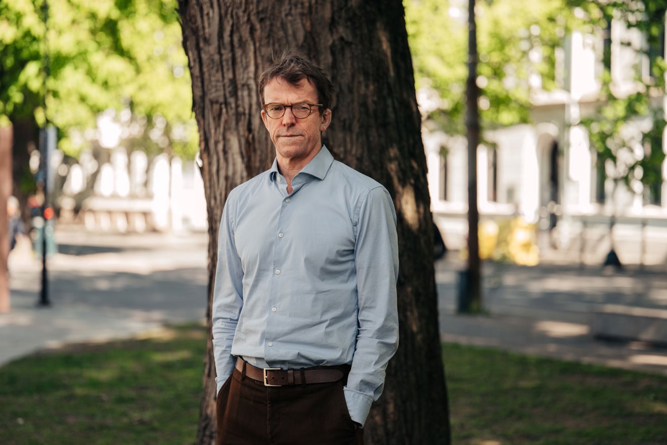 En voksen mann med briller står utendørs i blå skjorte foran et tre og grønne omgivelser ute.