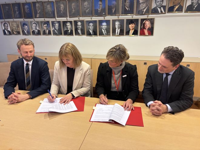 To kvinner sitter ved et bord og signerer dokumenter, med to menn i dress sittende på hver side.