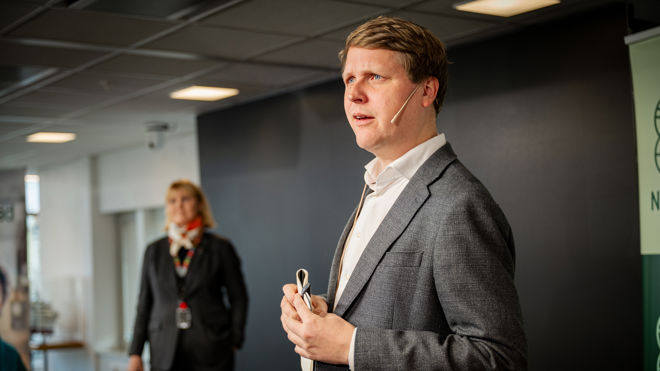 En mann i dress står foran og holder en mikrofon, mens en kvinne i dress står i bakgrunnen i et møterom.