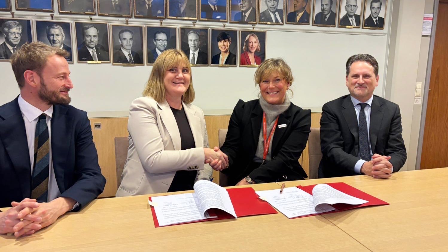 To kvinner sitter ved et bord og håndhilser foran dokumenter, med to menn i dress sittende på hver side.