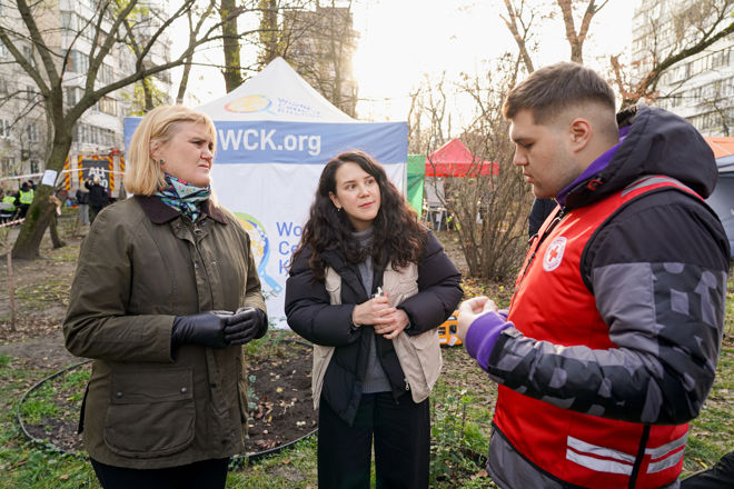 To kvinner og en mann står og snakker sammen utendørs foran et telt med World Central Kitchen-logo, og mannen har på seg en vest med Røde Kors-logo.
