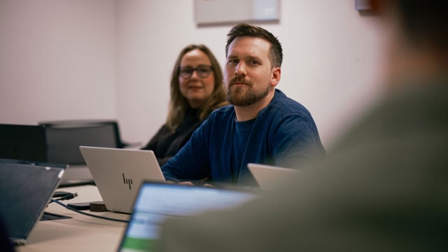 Two young adults sit at a meeting table with open laptops, while another person is partially visible in the foreground; a framed picture hangs on the wall behind them.