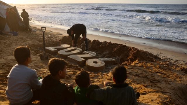 Fire barn sitter på stranden og ser mot en voksen som bygger store sandfigurer formet som tallet 2024 nær vannkanten.